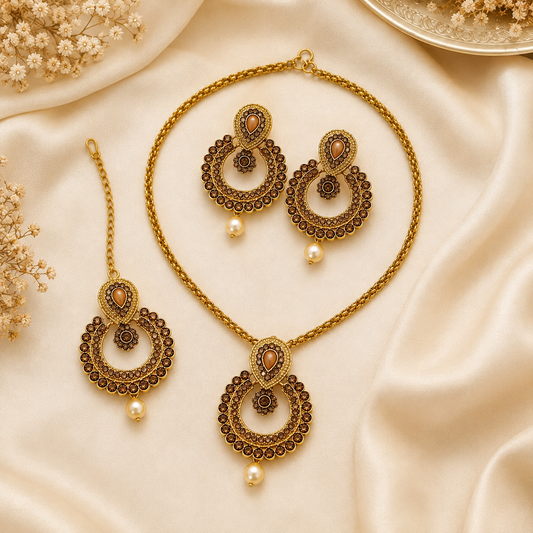 Gold and brown traditional Indian jewelry set including a necklace, matching earrings, and a maang tikka, arranged on a cream satin fabric background with dried floral decorations.