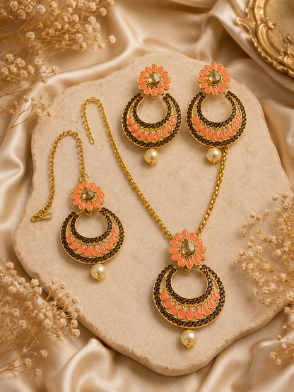 Gold-toned traditional jewelry set including a necklace, earrings, and a matching bracelet, decorated with coral-pink floral elements and dark metallic detailing, arranged on a beige stone slab with satin fabric and dried flowers.