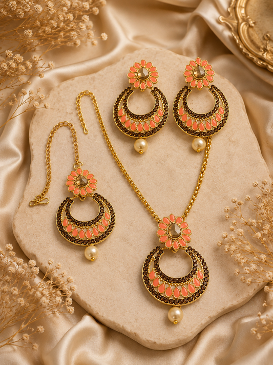 Gold-toned traditional jewelry set including a necklace, earrings, and a matching bracelet, decorated with coral-pink floral elements and dark metallic detailing, arranged on a beige stone slab with satin fabric and dried flowers.