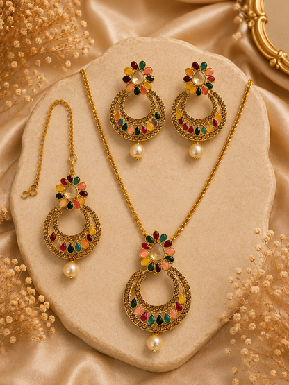 Gold-toned jewelry set with a necklace, earrings, and matching pendant displayed on a beige stone slab. The pieces feature ornate circular motifs with multicolored enamel-like floral accents in red, green, yellow, and pink, plus small pearl-like drops, set against a champagne satin background with decorative dried flowers.