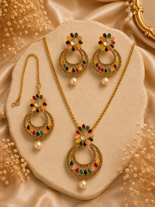 Gold-toned jewelry set with a necklace, earrings, and matching pendant displayed on a beige stone slab. The pieces feature ornate circular motifs with multicolored enamel-like floral accents in red, green, yellow, and pink, plus small pearl-like drops, set against a champagne satin background with decorative dried flowers.