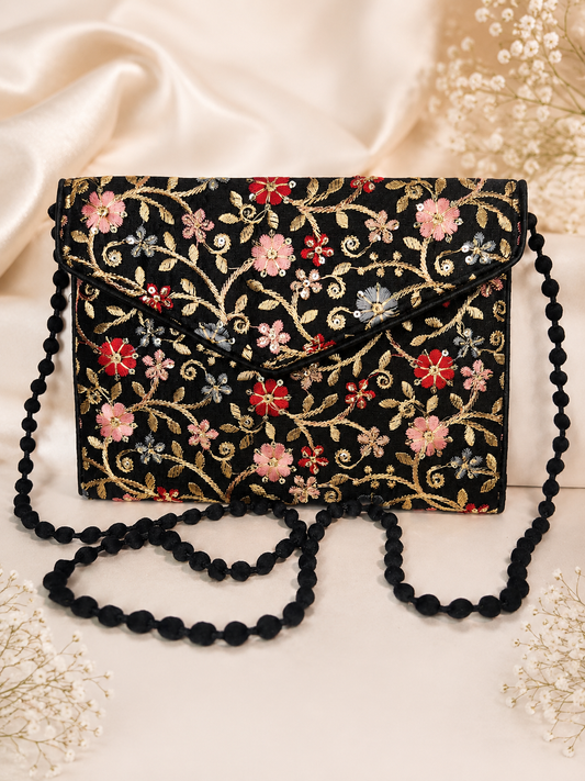 A black clutch bag with intricate floral embroidery in pink, red, and gold, featuring a black beaded shoulder strap, styled on a cream satin fabric background with dried flowers.