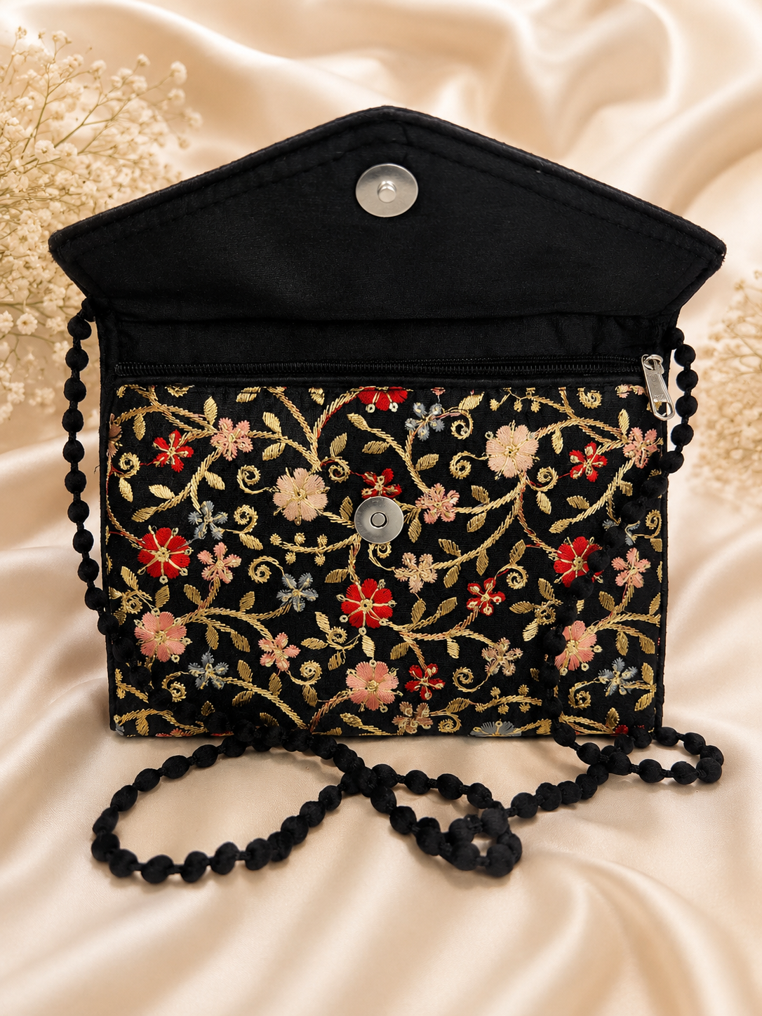A black fabric handbag with a pointed flap, decorated with colorful floral embroidery in red, pink, and gold on a black background. It features a black beaded shoulder strap and a silver snap button closure, displayed on a beige satin fabric background.