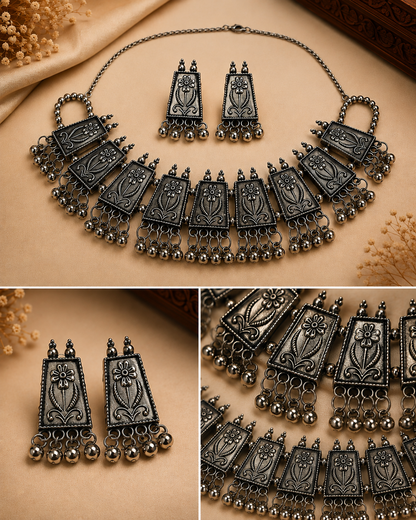 A traditional Indian-style jewelry set featuring a statement necklace, matching earrings, and a small pendant, all in an antique silver finish with floral engravings and dangling bell chains. The set is displayed on a beige fabric background with decorative dried flowers.
