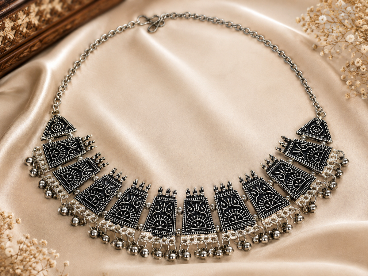 A silver-toned statement bib necklace with intricate black filigree patterns and dangling bead chains, displayed on a beige satin fabric background with decorative dried flowers.