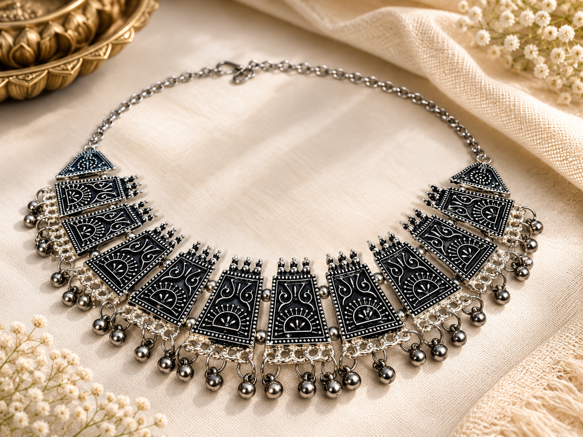 A silver-toned statement collar necklace with black enamel panels, intricate filigree details, and dangling silver beads, displayed on a cream fabric background with decorative dried flowers.