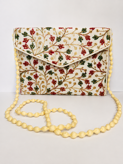 An embroidered cream clutch bag with a floral vine pattern in red, green, and gold, featuring a matching cream beaded shoulder strap.