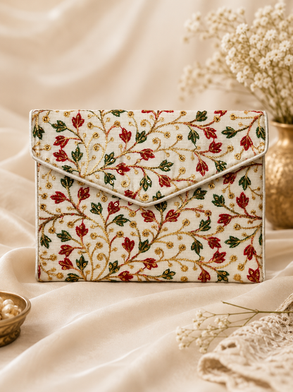 A cream-colored clutch bag with an intricate floral vine pattern in red, green, and gold embroidery, styled on draped cream fabric with dried flowers in the background.