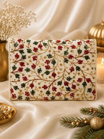 An embroidered clutch bag with a cream background and a festive pattern of red and green leaves, gold vine details, and small gold dots, styled on draped satin fabric with holiday decorations.