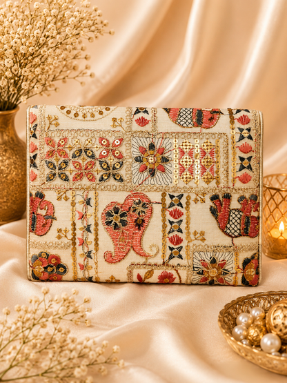 An embroidered clutch bag with a patchwork design featuring floral, geometric, and elephant motifs in red, gold, and black on a cream background, styled on draped satin fabric with dried flowers and a candle.
