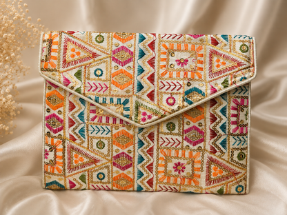 A rectangular clutch bag with a vibrant, multicolored geometric and tribal-inspired embroidery pattern on a cream base, styled on a draped beige satin fabric background.