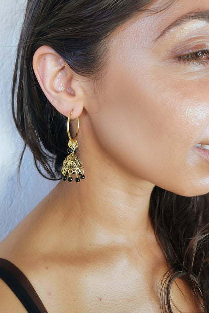 A close-up of a woman's ear wearing a gold huggie hoop earring with a dangling ornate filigree jhumka-style charm and small black bead drops.