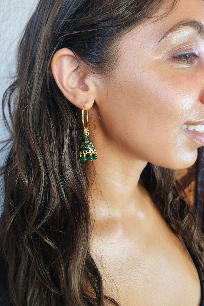 A close-up side profile of a woman wearing a gold huggie hoop earring with a dangling green and gold ornate charm.