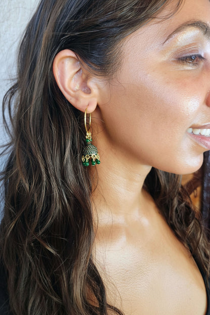 A close-up side profile of a woman wearing a gold huggie hoop earring with a dangling green and gold ornate charm.