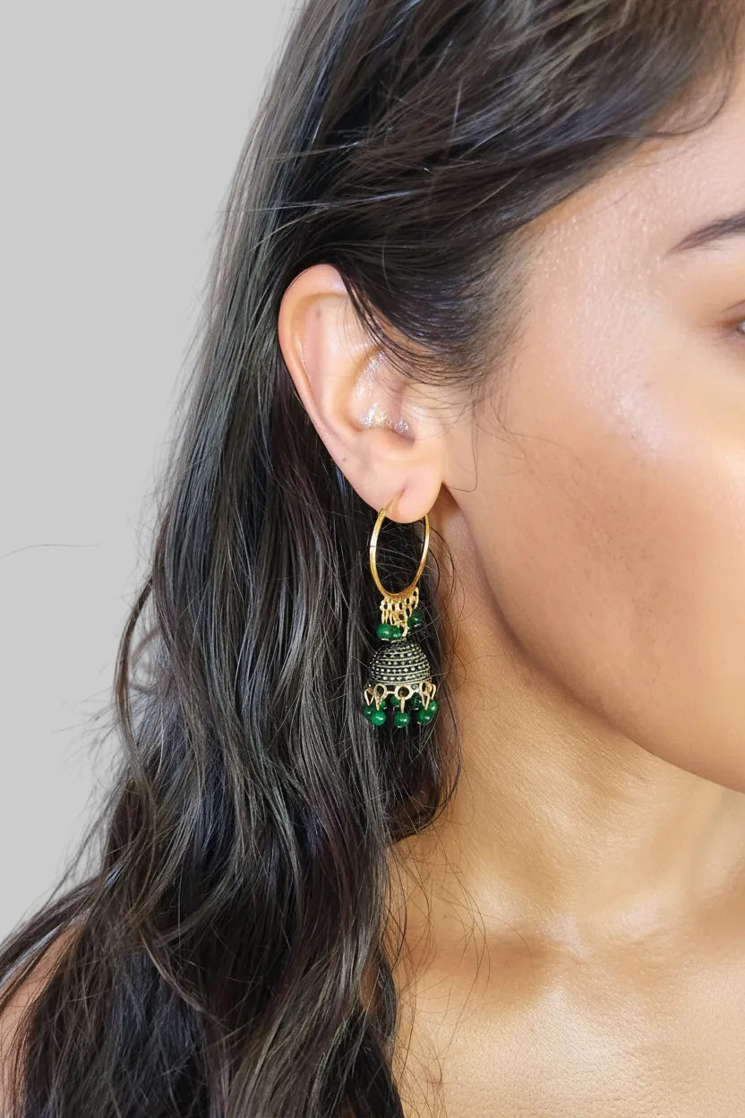 A close-up of a woman's ear wearing a gold hoop earring with a dangling bell-shaped charm embellished with green stones.