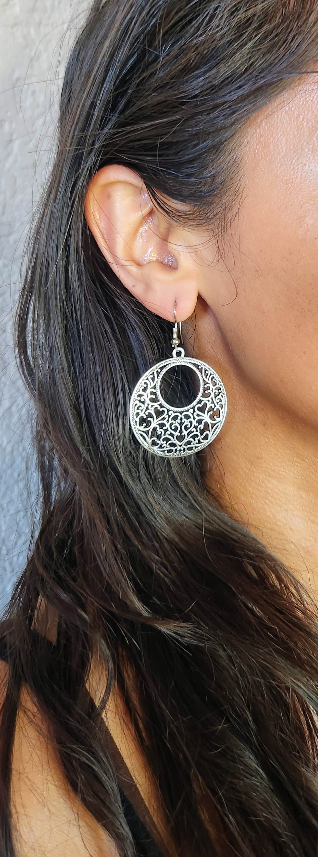 A close-up of a woman's ear wearing a silver filigree circular drop earring with a black center, against a neutral gray background.