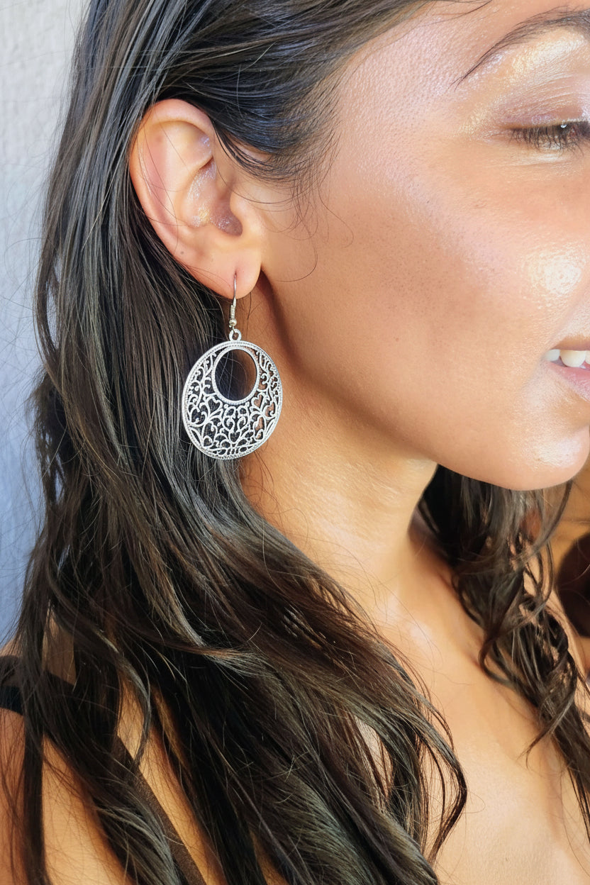 A close-up of a woman wearing a silver filigree dangle earring with a dark oval center, shown against her ear and dark hair.