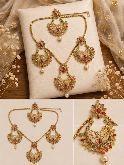 Gold-toned traditional Indian jewelry set with a necklace, earrings, and maang tikka, decorated with red stones, green stones, and white pearl beads, displayed on a cream cushion and fabric background.