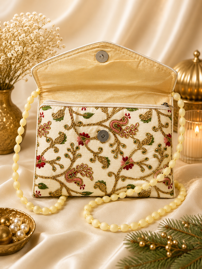 An embroidered clutch handbag with a cream base, detailed with gold paisley and floral embroidery, red floral accents, and a pearl-bead strap, styled on a draped satin fabric with decorative props.