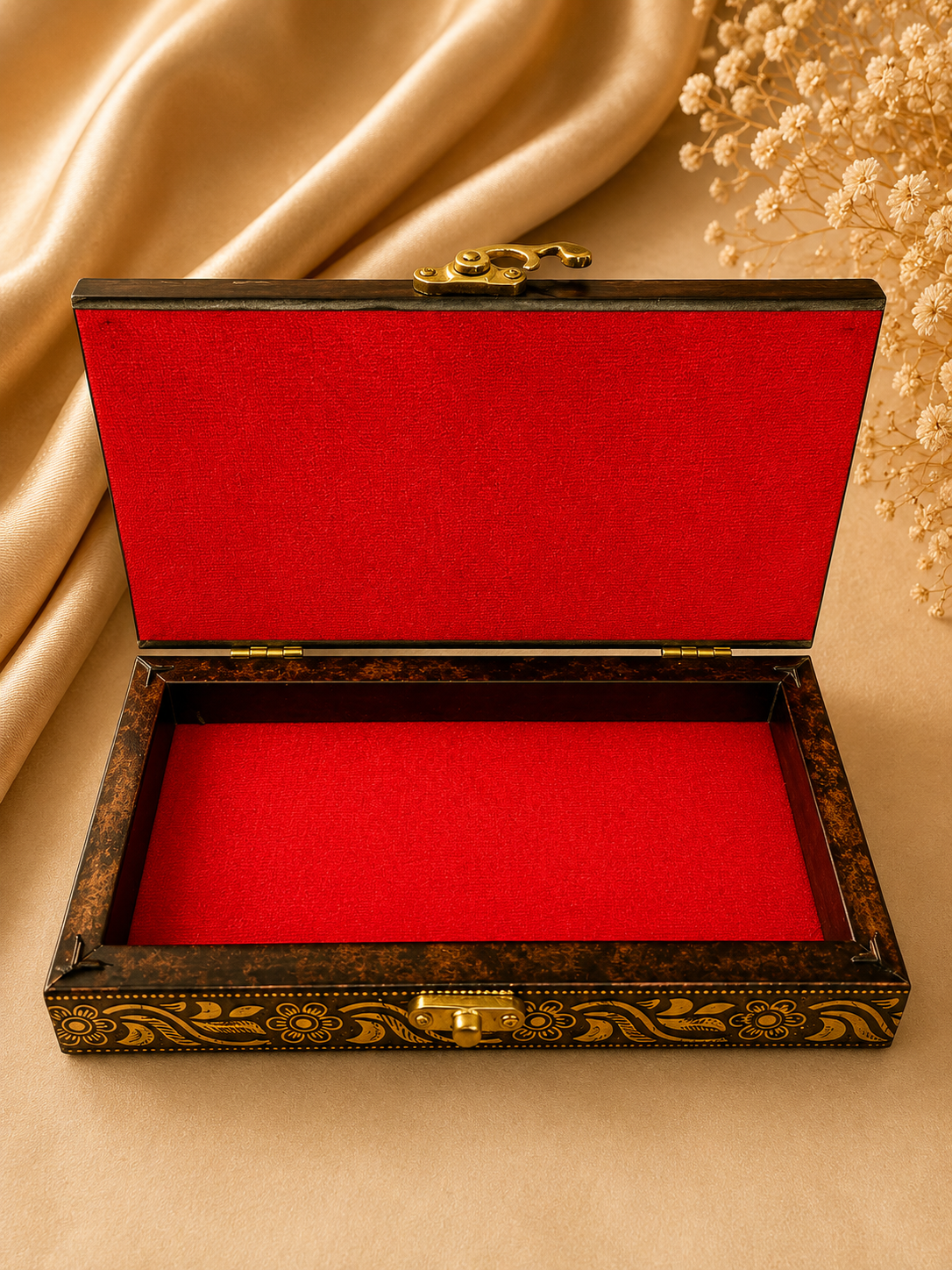 An open rectangular jewelry box with a dark brown marbled wood exterior, gold decorative scrollwork, and a bright red velvet interior, styled on a beige fabric background with draped satin and dried flowers.