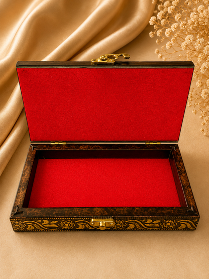 An open rectangular jewelry box with a dark brown marbled wood exterior, gold decorative scrollwork, and a bright red velvet interior, styled on a beige fabric background with draped satin and dried flowers.