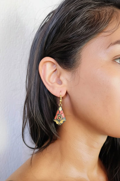 A close-up of a woman wearing a dangling earring. The earring has a gold-toned metal frame with a colorful floral or lotus-like design in red, green, yellow, and white enamel.