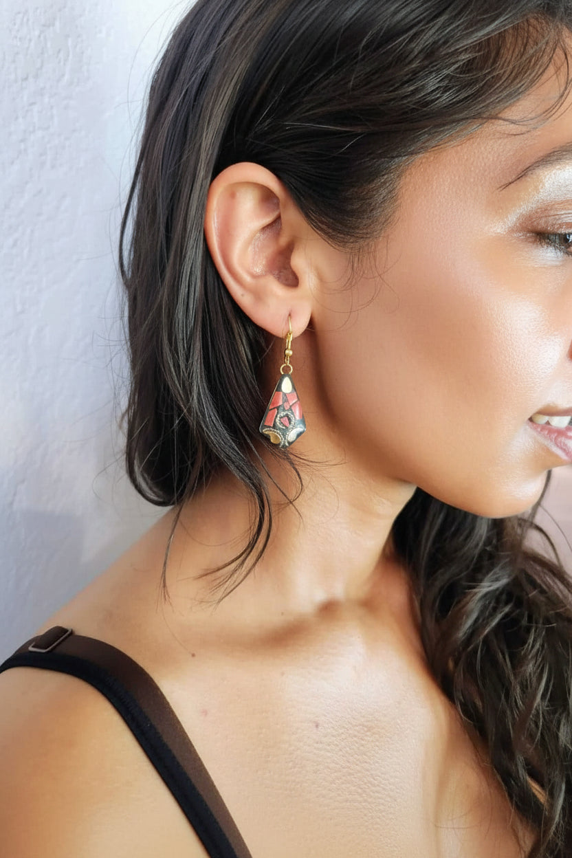 A close-up side profile of a woman wearing a dangling earring. The earring has a gold-toned hook and a colorful, hand-painted triangular pendant with pink, blue, and green details.