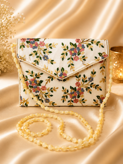 A cream-colored fabric clutch bag with an embroidered floral pattern in pink, blue, and green, featuring a matching cream flap and a long cream beaded shoulder strap, displayed on a draped satin background.