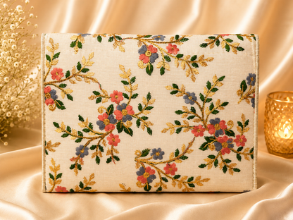 A rectangular clutch purse with an embroidered floral pattern on a cream linen base, styled on draped champagne satin with a gold candle holder and dried flowers.