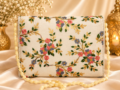 A rectangular cream-colored clutch bag with an intricate floral embroidery pattern in pink, blue, green, and gold. The bag features a cream beaded strap and is styled on a draped satin fabric with decorative gold-toned props in the background.